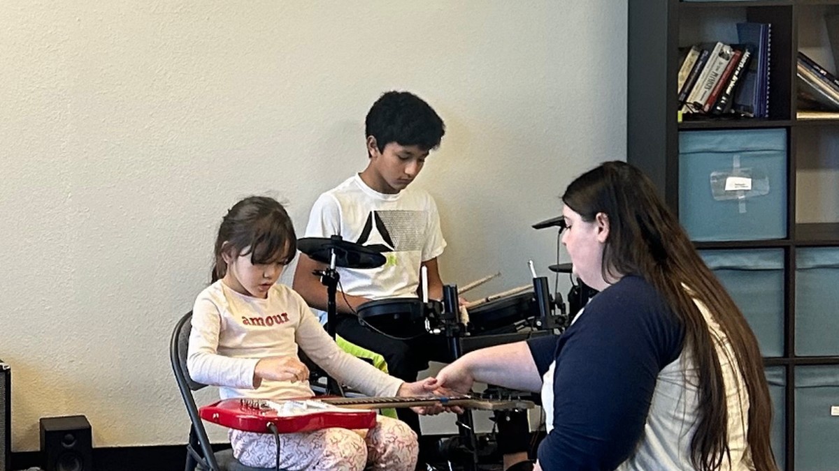 Rising Rockers instructor helping a young student with a red electric guitar while another student plays drums